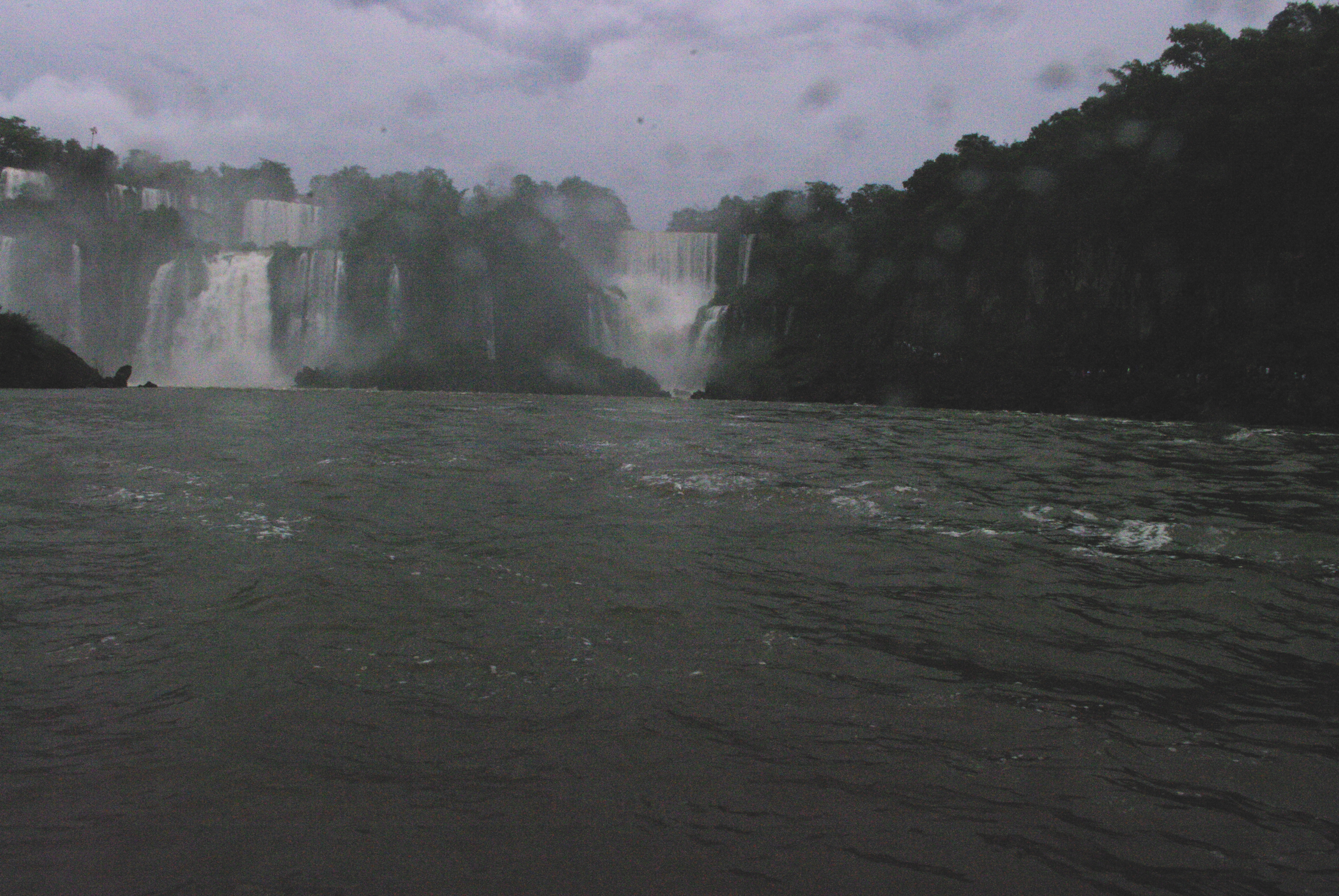 On a boat trip on the lower river, with the green forest on the side of the blueish river. We are now approaching two of the smaller waterfalls, separated by a small forested island.