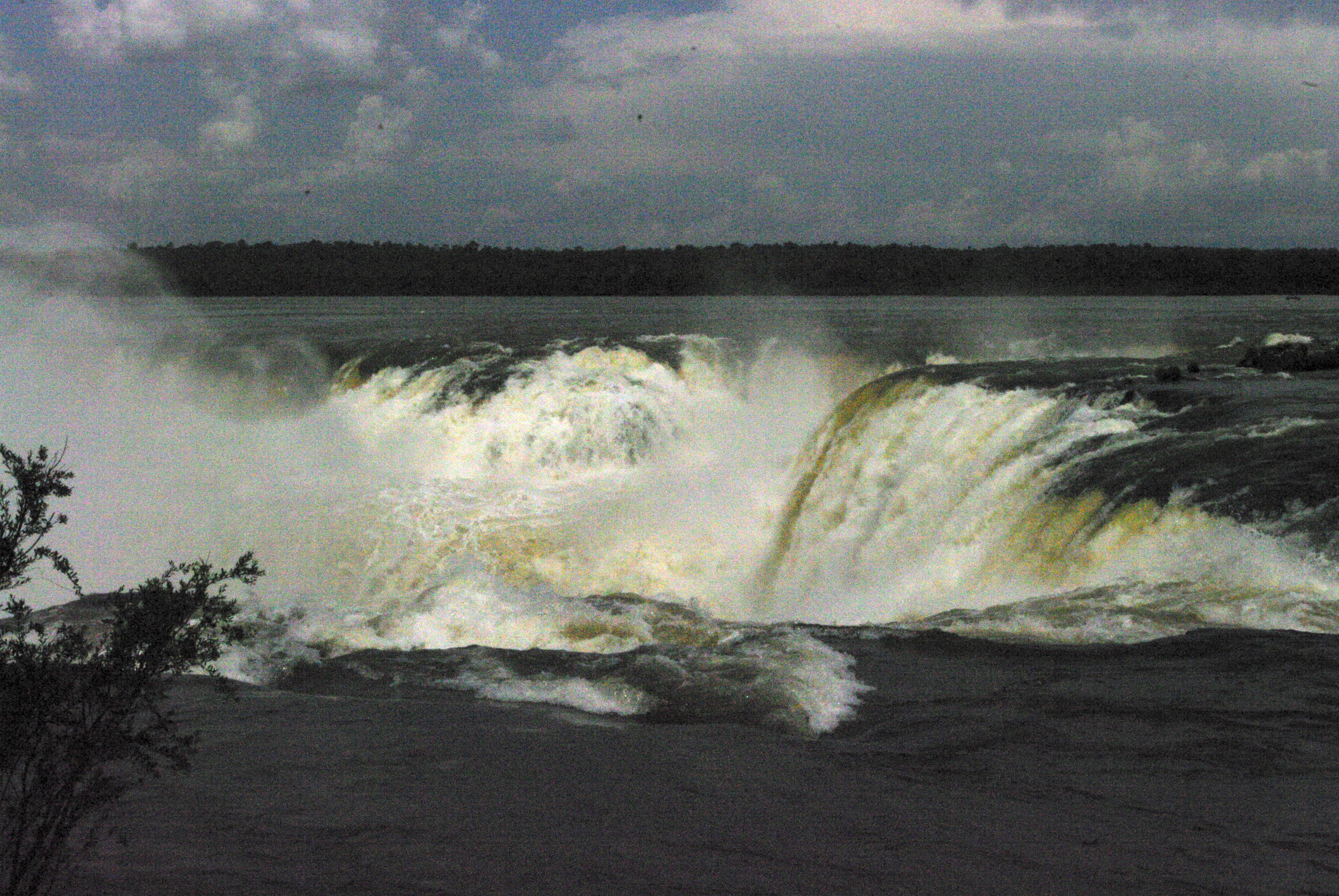 Looking across the river and Iguazu Falls from a viewing platform . You can see the water as it pours over the edge and heads downwards, producing large white clouds of mist above.