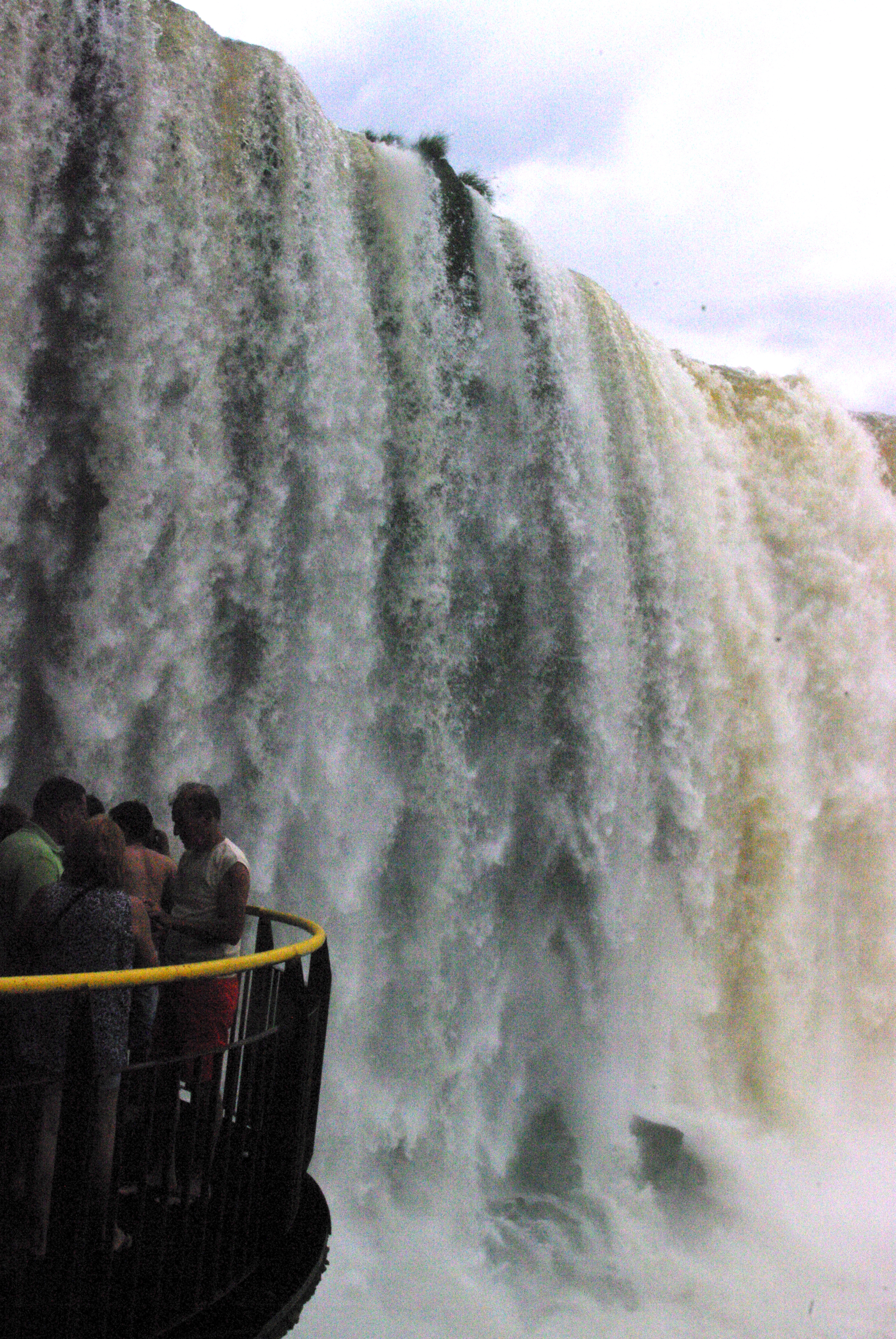 A view of one of the viewing platforms at the side of one of Iguazu's waterfalls, with some people standing looking at the plummeting water.