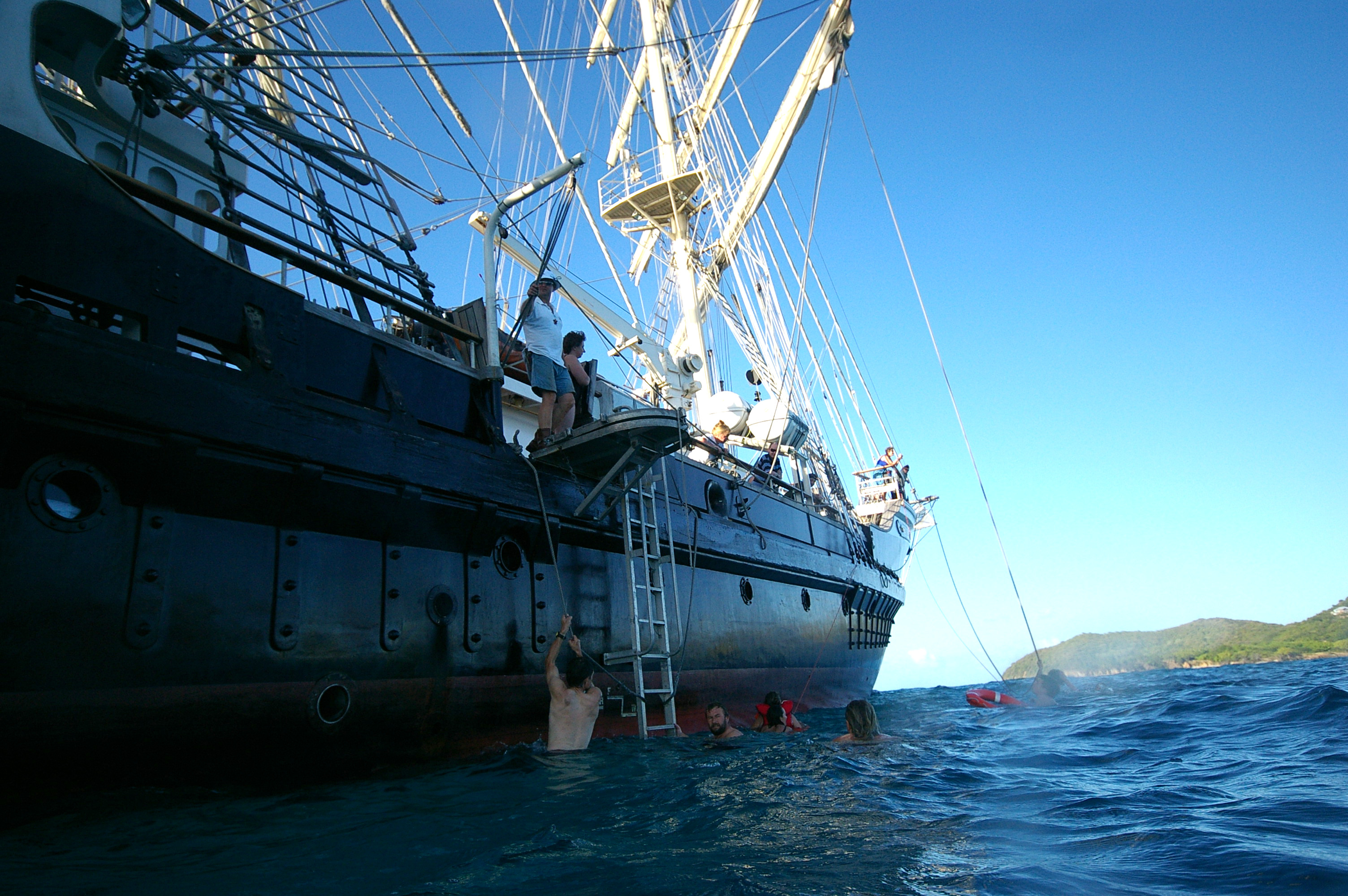 The bow of Tenacious, taken from the water,  anchored off Dominica. The island can be seen in the background, with a couple of the swimmers cdlimbing down into the water.