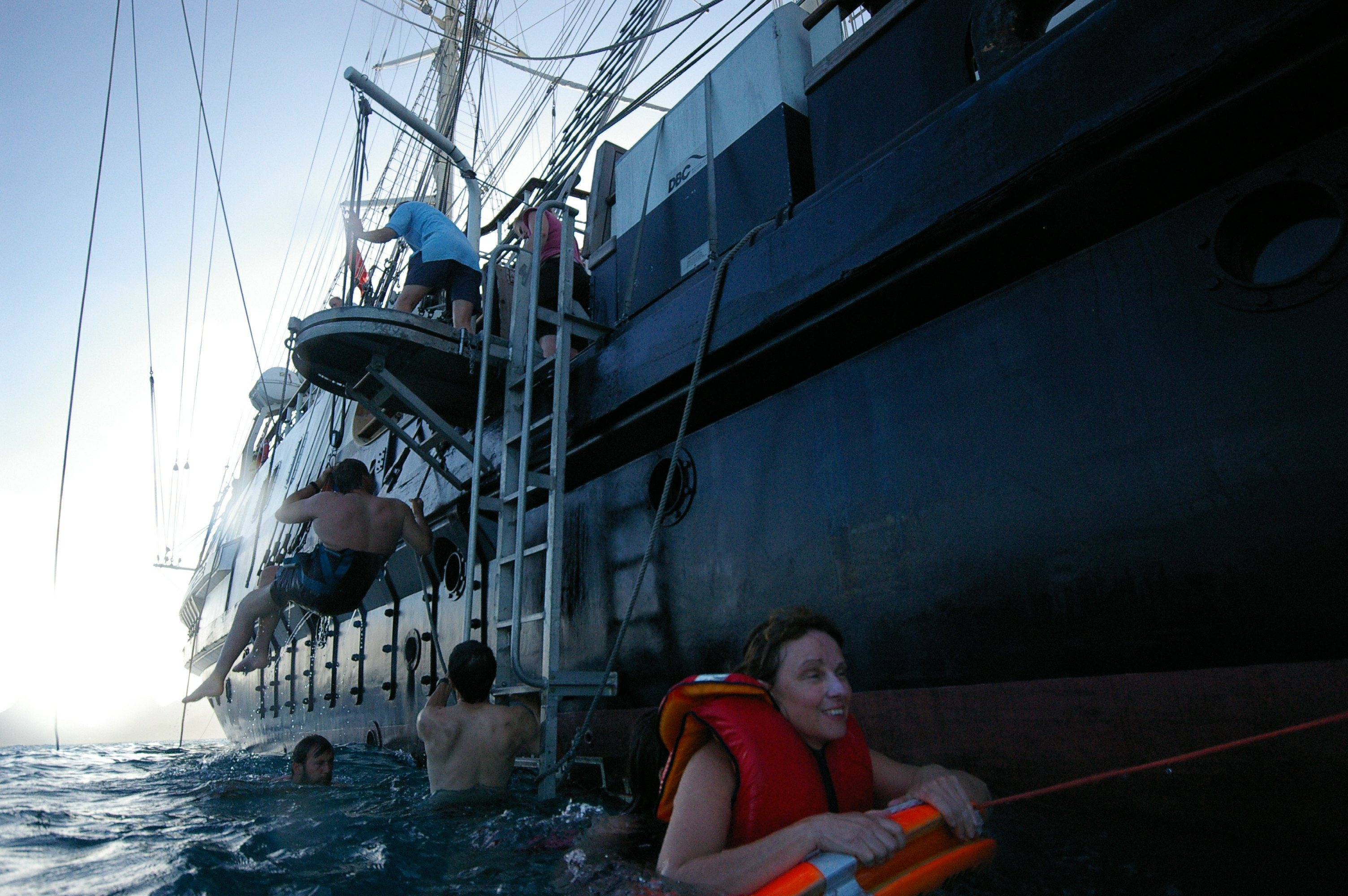 A picture taken from the water of a woman wearing a life jacket and using one of the life rings, another being lowered using the hoist and a third entering the water via a ladder